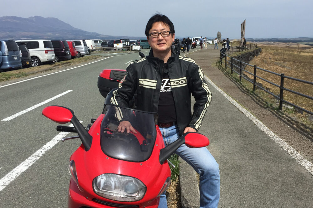 阿蘇の景色を背景に、赤いバイクを路肩に停めて、ライダージャケットを着て写真におさまる三橋先生